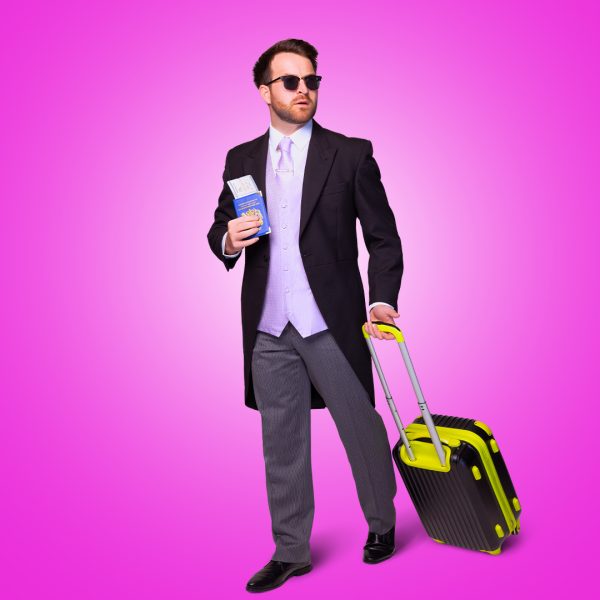 Billy Roberts as Nathan. He wears a black grooms suit with lilac waistcoat and tie. He has ray-ban sunglasses on and holds a passport and suitcase.