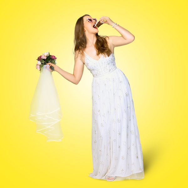 Lucie Mae-Sumner pictured against a bright yellow background in a wedding dress, holding a bouquet and veil in her right hand and eating a slice of cake in her left. She also wears heart sunglasses.