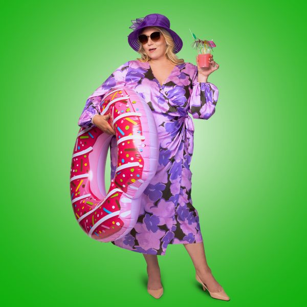 Melissa Jacques looks glamorous in a purple floral wrap dress. She is wearing dark purple sunglasses and a purple wedding hat. She holds a pink doughnut pool float and a fruit cocktail.