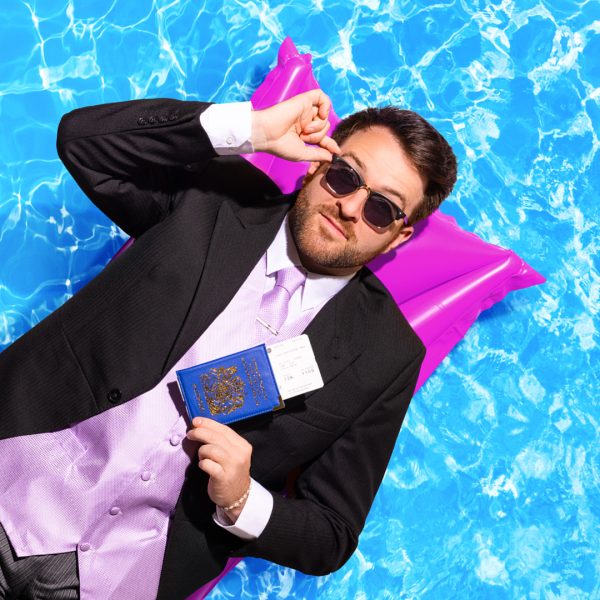 Billy Roberts as Nathan. He lies on a purple lilo against a pool backdrop. He has a suit on, sunglasses and holds a passport.
