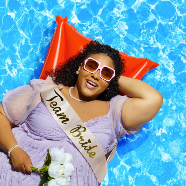 Kayla Carter as Bonnie laying on a lilo with a pool backdrop. She wears lilac bridesmaid dress and a Team Bride sash.