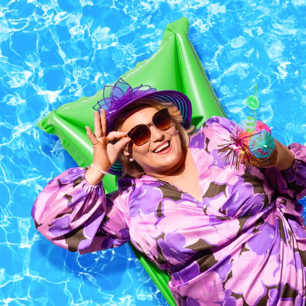 Melissa Jacques pictured in a purple floral dress, wearing a purple wedding hat and dark sunglasses. She lies on a green lilo with a pool water backdrop. She pinches her sunglasses between two fingers and holds a cocktail in the other hand.