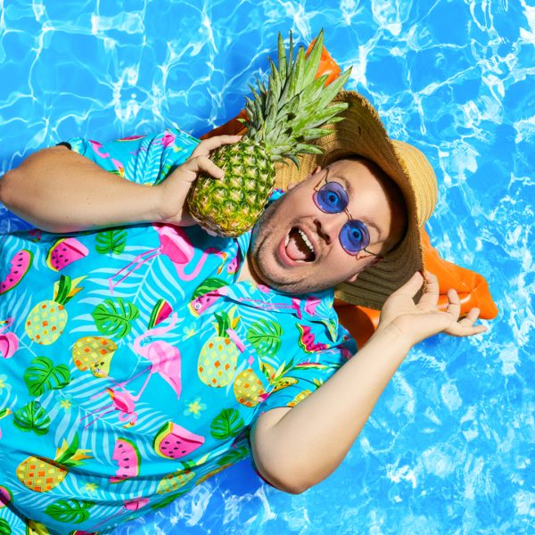 Cast image of Scott Paige. Scott is wearing a Hawaiian shirt, blue sunglasses and a straw hat. He is lying on a lilo with pool water as the background. He's holding a pineapple to his ear and looks ecstatic about it.