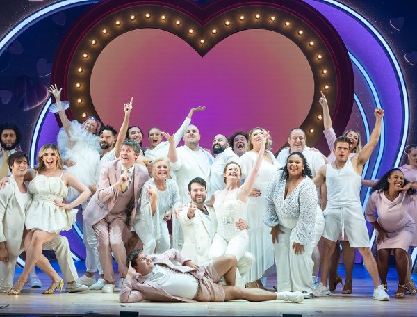 Close up of ensemble cast of I Should Be So Lucky the musical all dressed in white. A purple-pink love heart is in the background with gold lights around it. The cast are reaching to the sky as well as pointing out to the audience.