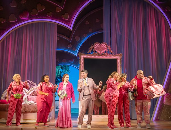 A line of the I Should Be So Lucky ensemble stand at the front of the stage. In the background is a staggered heart design with a huge pink ornate mirror in the centre. A pink heart shaped bed is to the left of the stage. The cast are all dressed in different eclectic pink outfits. A man stands in the middle in a dusty pink suit.