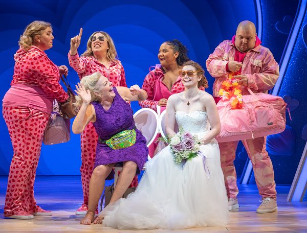 A smiling bride in her wedding dress, holding a white & purple bouquet & wearing pink heart sunglasses sits next to a smiling older woman in a purple dress with a green bumbag. Behind them are 3 women in pink heart print tracksuits. On the right end of the line is a man in a pink tie-dye outfit holding a pink bag & an orange lei.