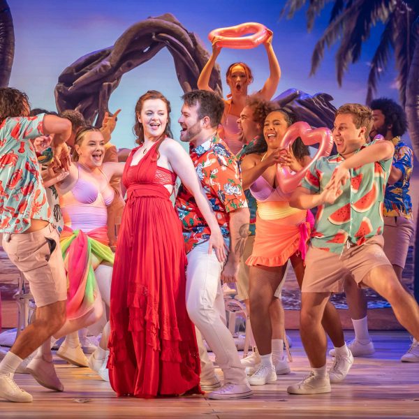 The cast of I Should Be So Lucky the musical on stage. A woman in a long red dress dances with a man in an orange Hawaiian shirt. They are surrounded by an ensemble of dancers; guys in Hawaiian shirts and girls in sarongs and swimsuits. Some carry pink inflatable love hearts. There's a tropical beach scene in the background.