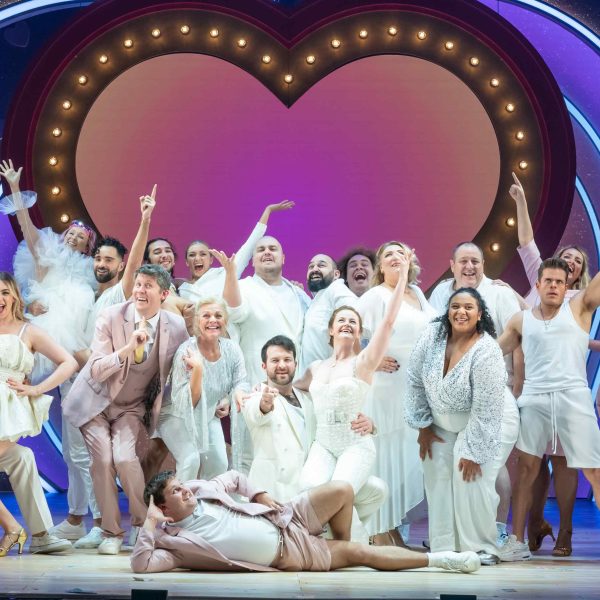 Close up of ensemble cast of I Should Be So Lucky the musical all dressed in white. A purple-pink love heart is in the background with gold lights around it. The cast are reaching to the sky as well as pointing out to the audience.
