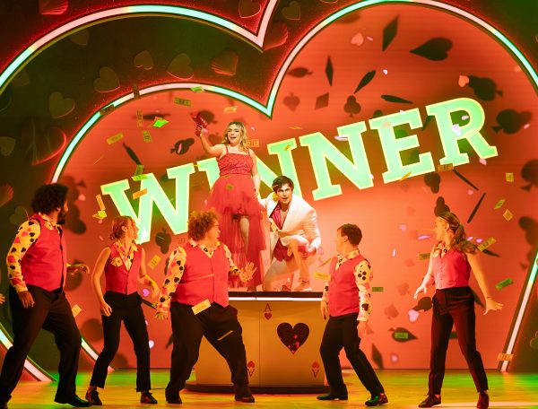 On-stage photo of I Should Be So Lucky the musical. A neon WINNER sign is in the background. A man & a woman stand on a table, the woman is holding a money shooter and is shooting bills across the stage. The man is wearing a white suit and is crouching on the table. Around them on the ground are 7 dancers wearing casino dealer outfits.