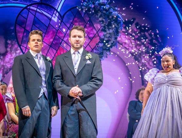 A wedding photo. A bridge (or bridesmaid) dressed in a frilly lilac dress looks left with her mouth open in a shocked smile. Two men in grooms suits with purple ties stand on the left looking confused. There is purple confetti in the air and a stained glass heart shaped window behind. Wedding guests sit behind the front 3 people.
